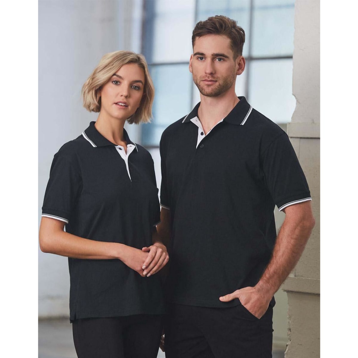 A unisex black polo shirt featuring white detailing, modelled by a man and woman in an indoor setting.