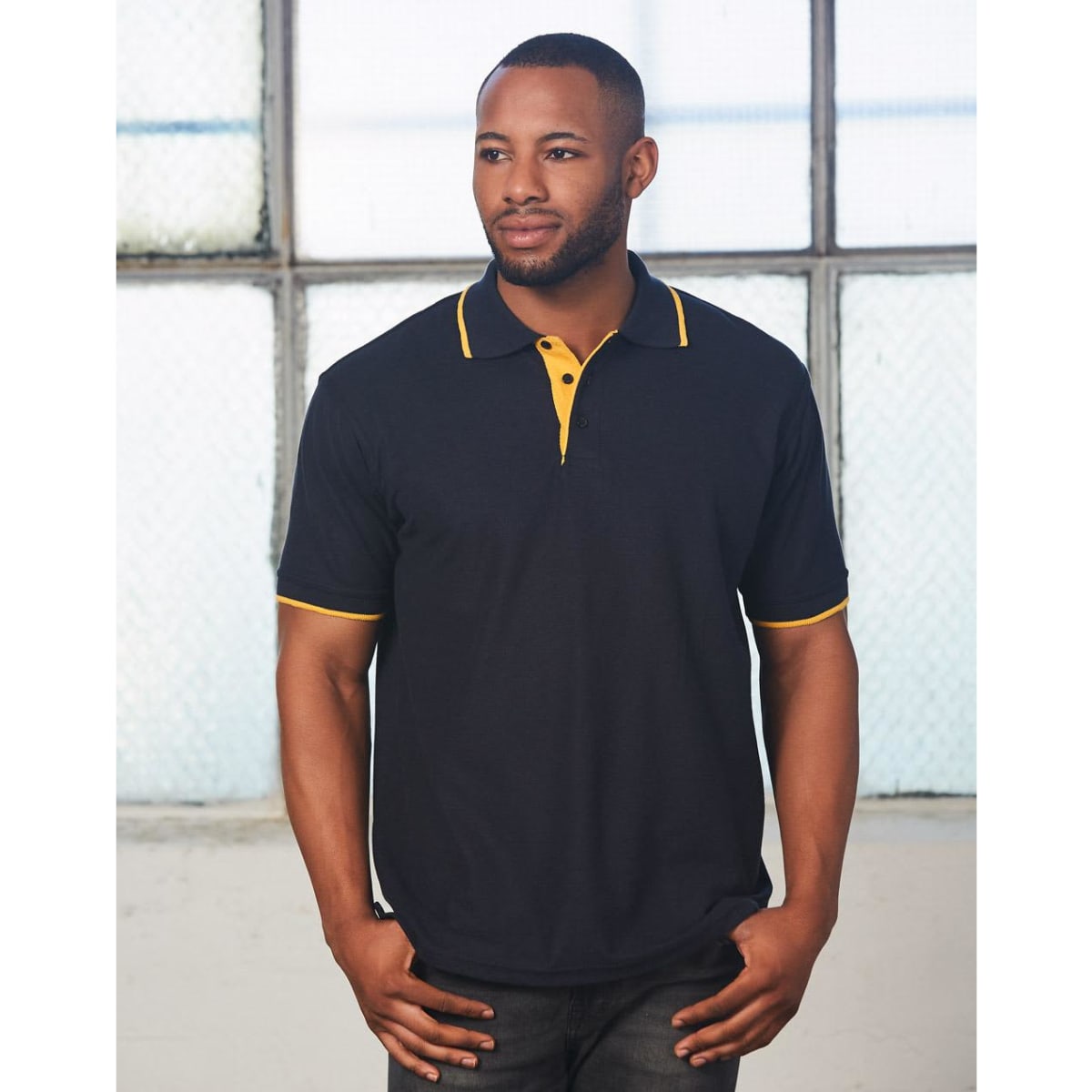 A black polo shirt with yellow detailing, featuring a collar and a logo, modeled by a man in an industrial setting.