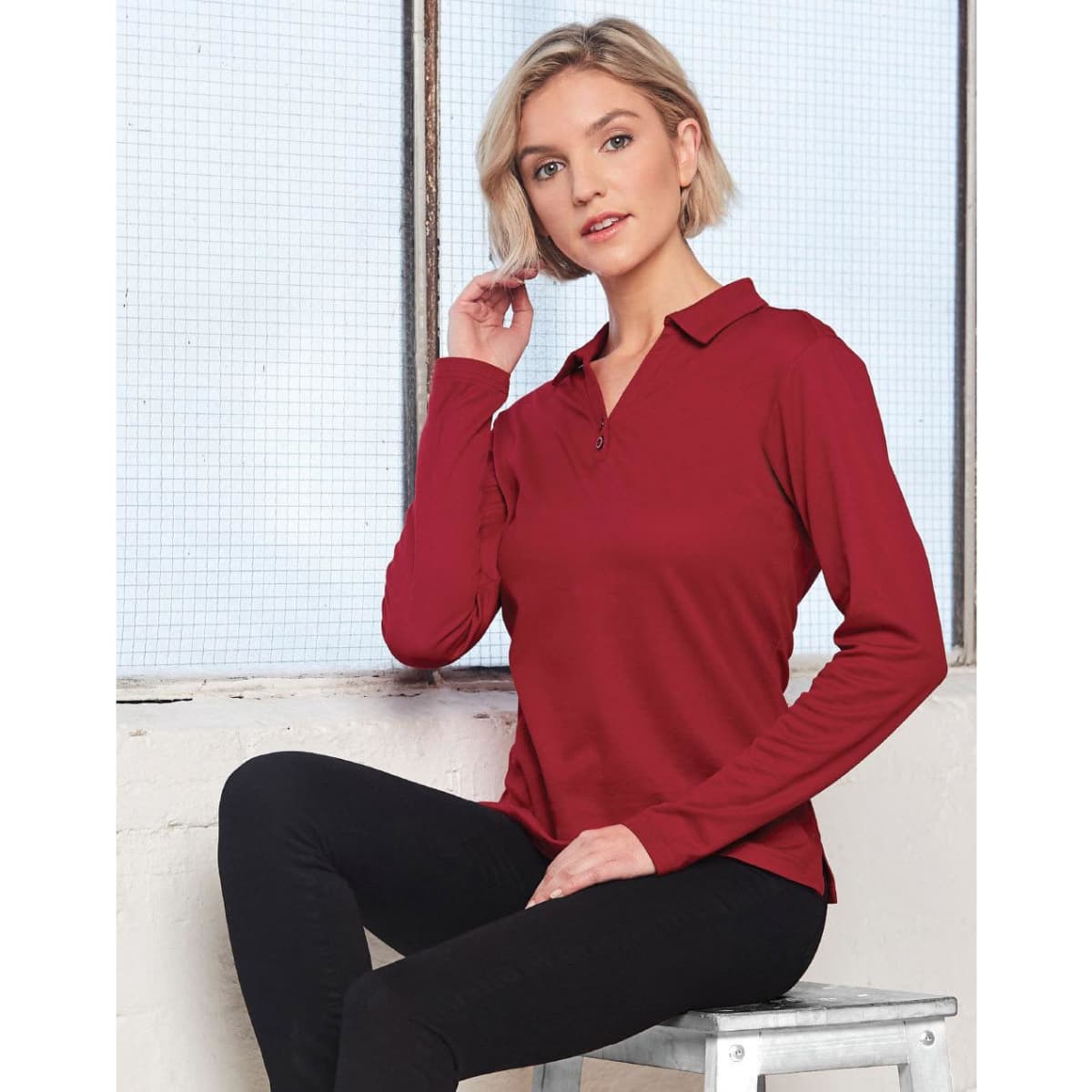 A long sleeve polo shirt in deep red, featuring a collar and a subtle logo. The model poses against a window.