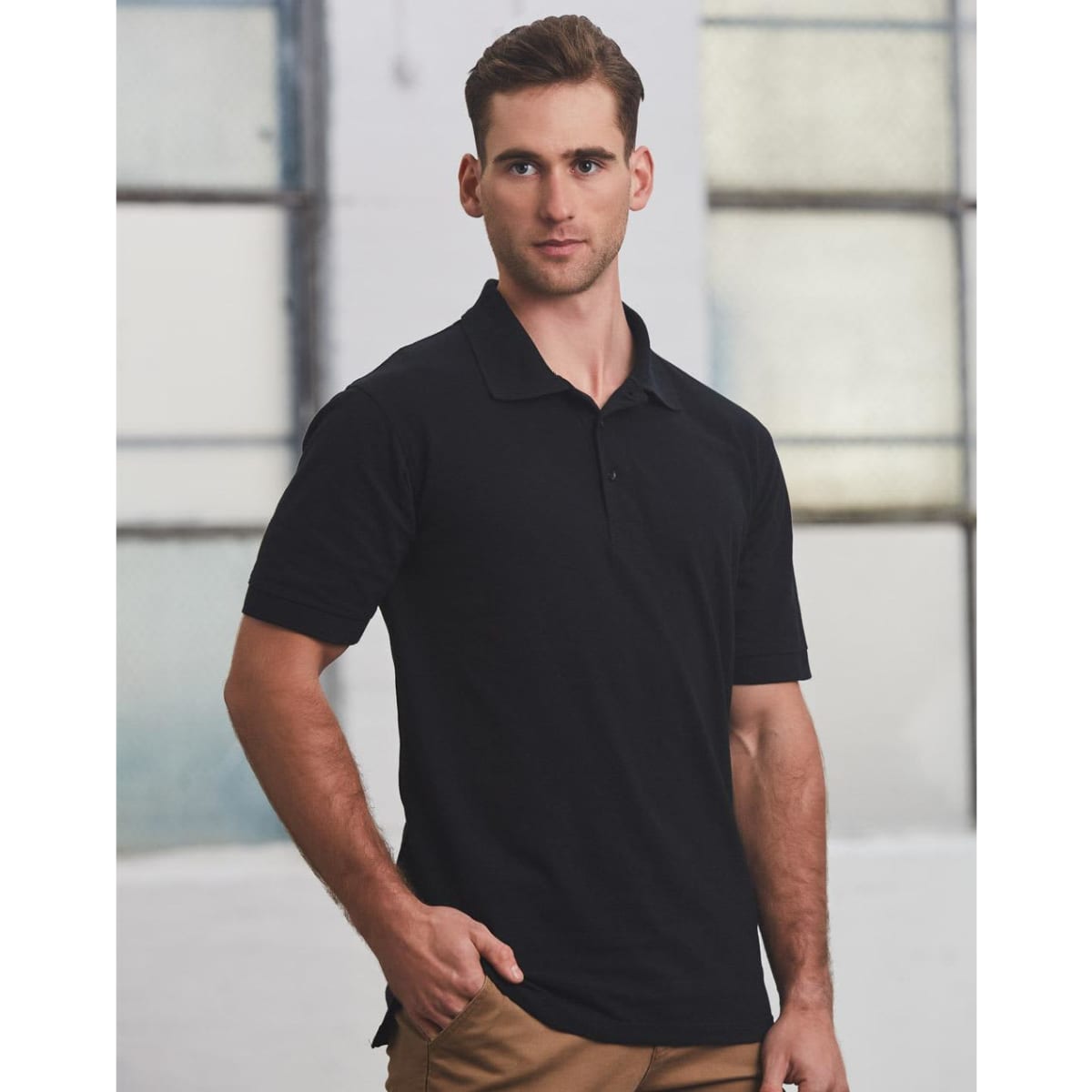 A man in a black polo shirt stands against an industrial background, featuring short sleeves and a standard collar.