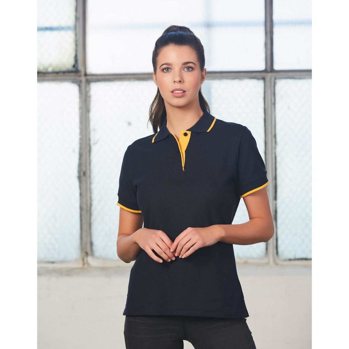 A black polo shirt with yellow accents, featuring a collar and a logo. The model stands in front of a window.