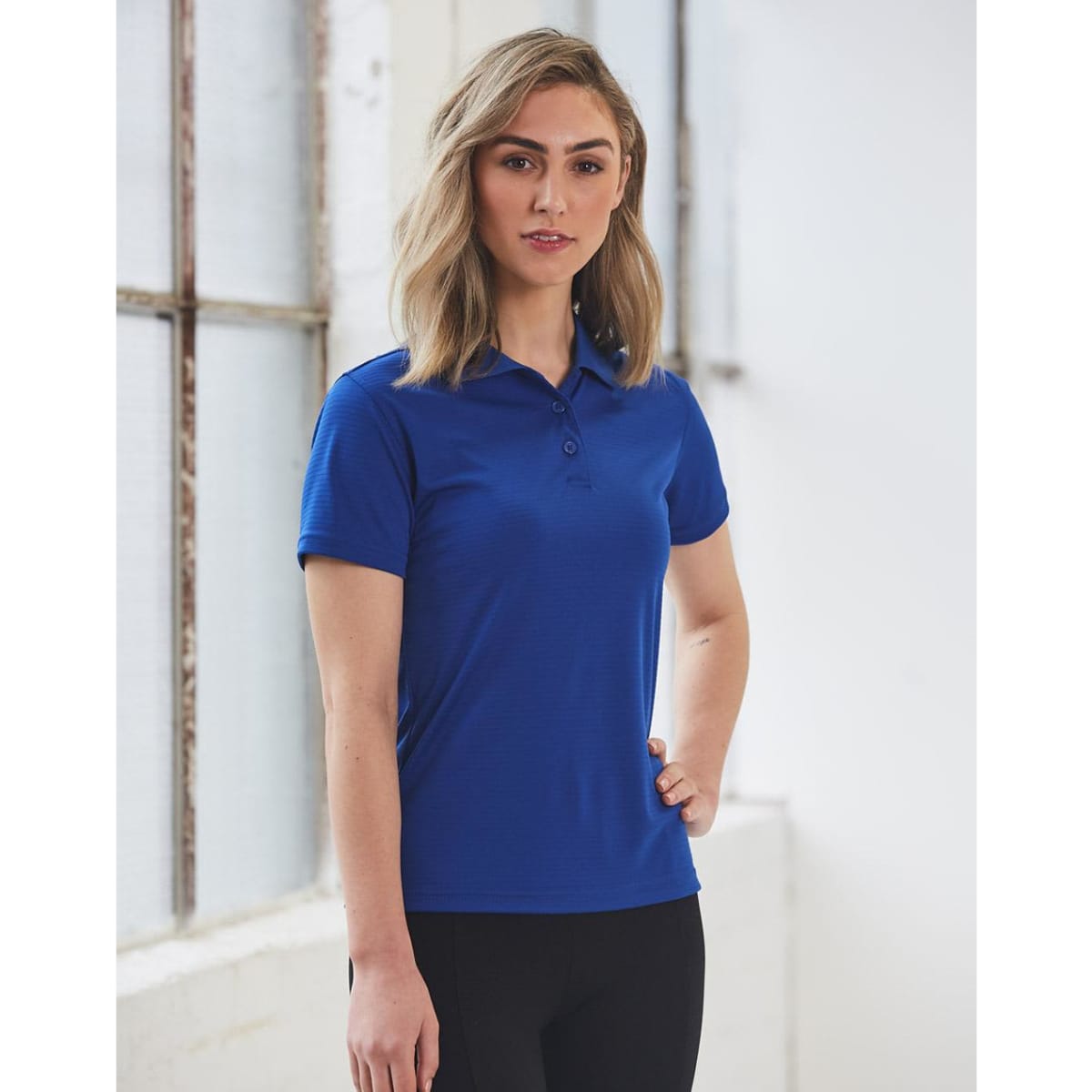 A woman stands wearing a ladies' textured polo shirt in blue, featuring a collar and a logo.