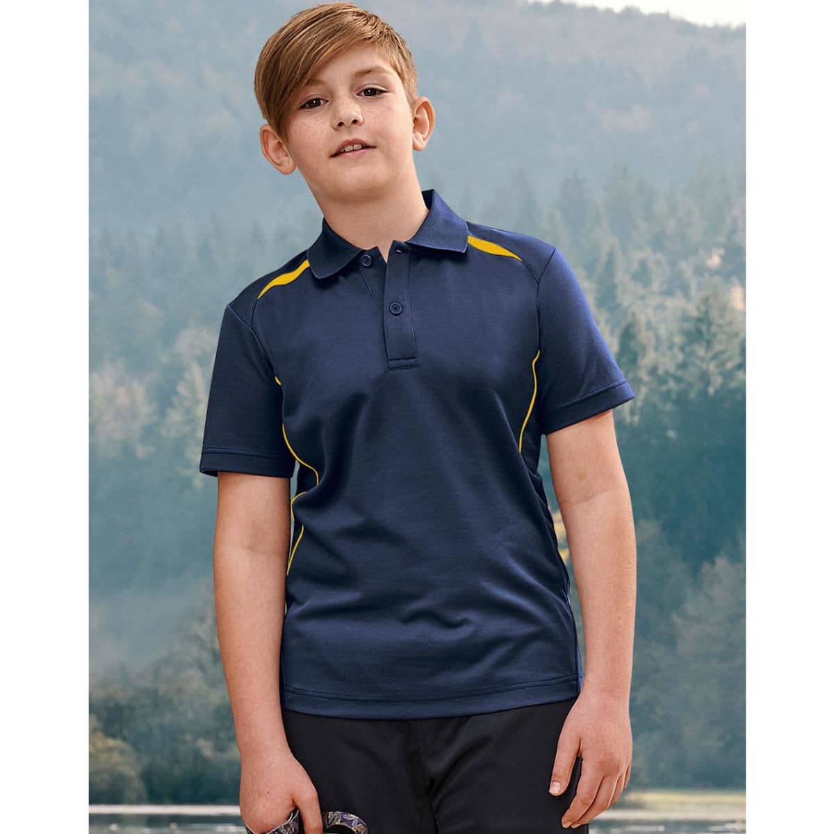 A boy stands outdoors wearing a navy polo shirt with yellow accents and a logo. The background features trees and hills.
