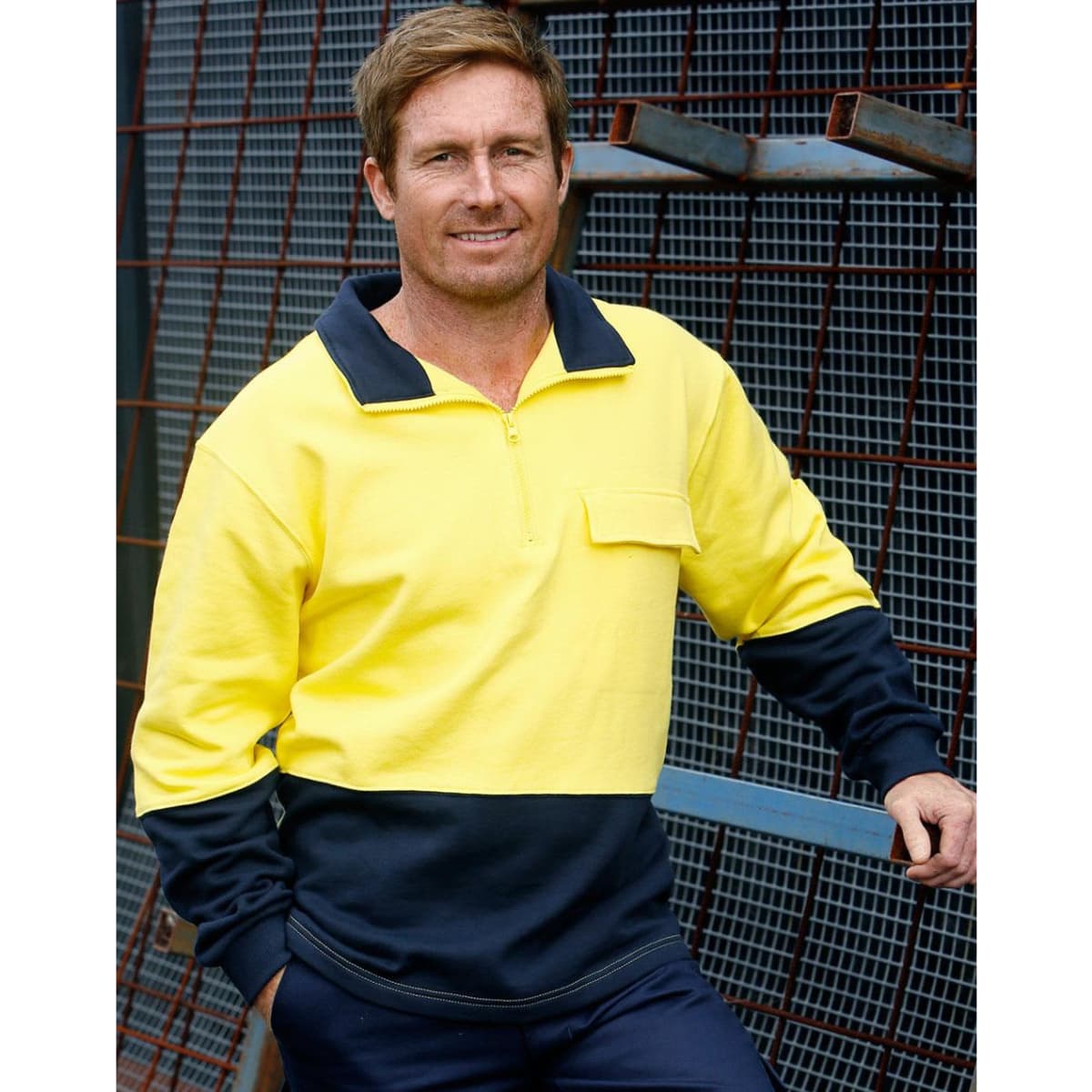 A man wearing a hi-vis two-tone yellow and navy cotton fleecy sweatshirt with a zip and pocket, posed against a metal backdrop.