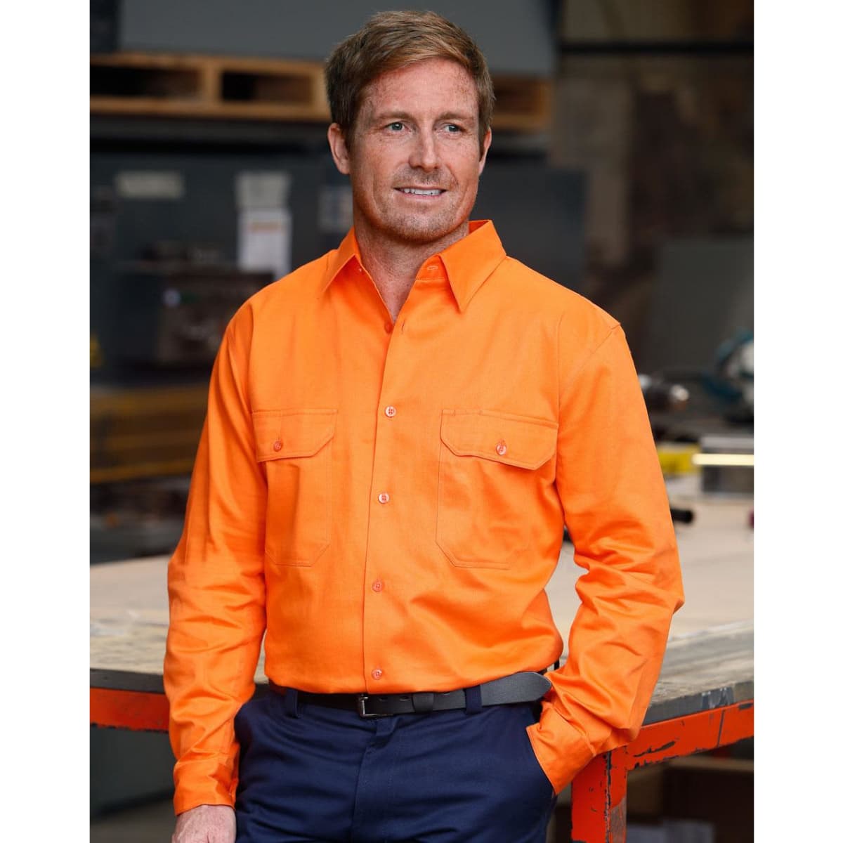 A bright orange men's long-sleeve drill shirt with two front pockets and a logo, set against an industrial background.
