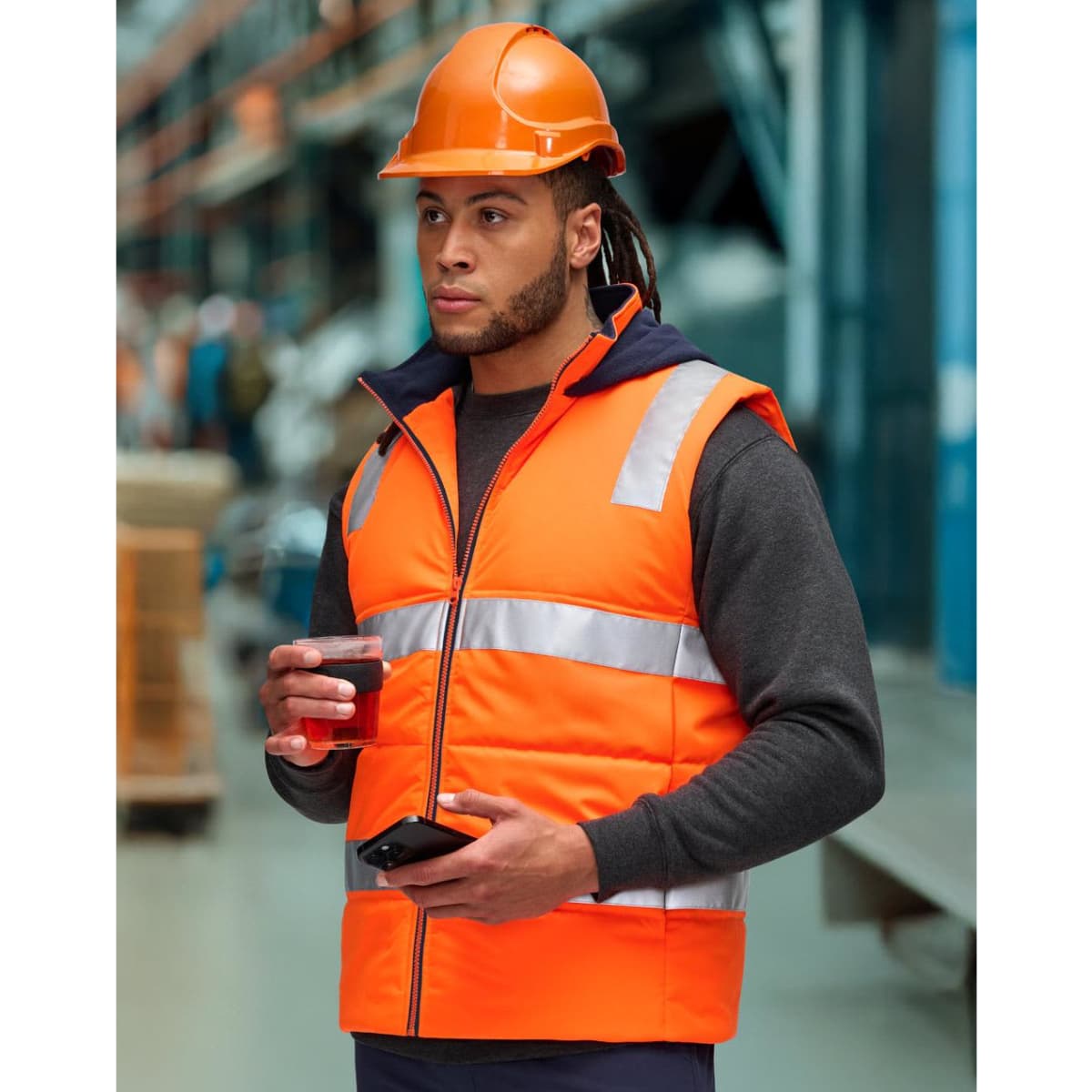 A man wearing an orange hi-vis hooded puffer vest and a hard hat stands in a warehouse, holding a coffee cup.