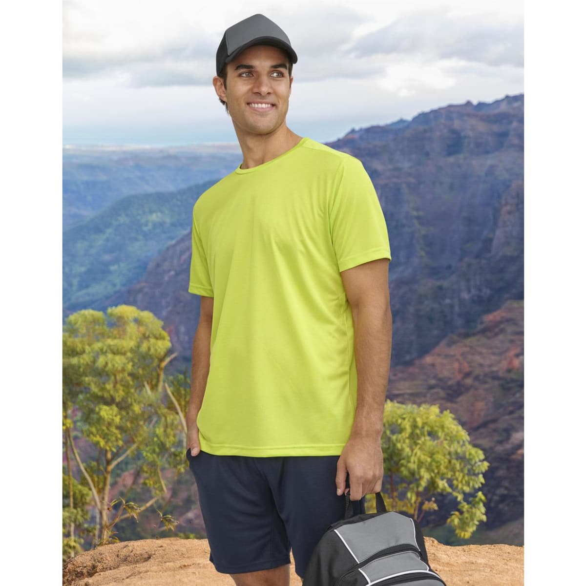 A man wearing a bright yellow short-sleeve performance T-shirt stands against a mountainous backdrop, holding a bag.