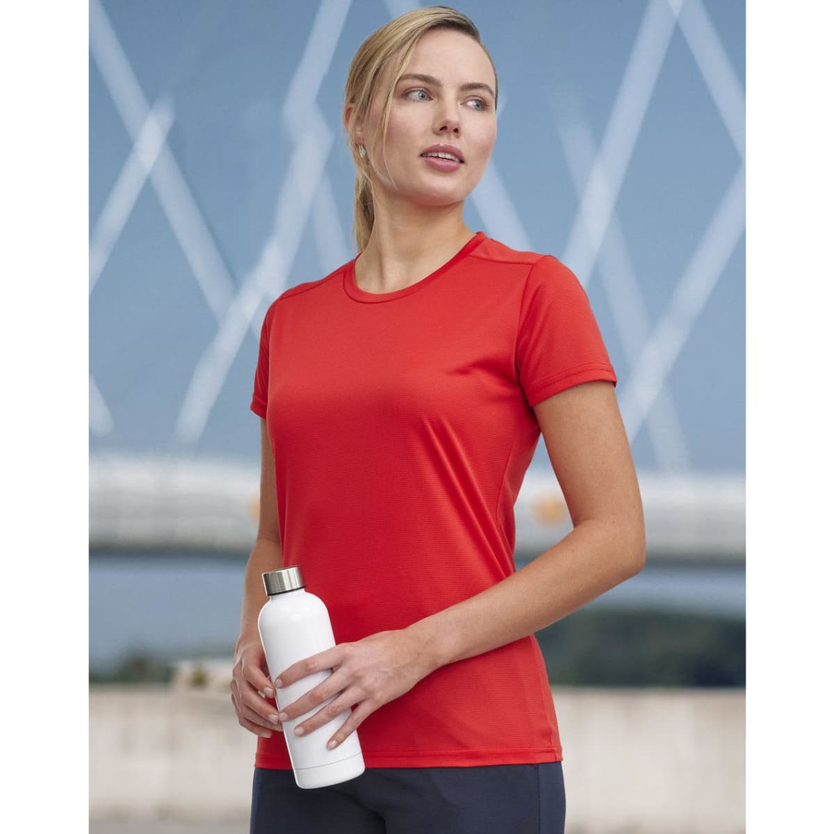 A woman wearing a red short-sleeved performance t-shirt holds a white drink bottle, set against a blurred outdoor background.