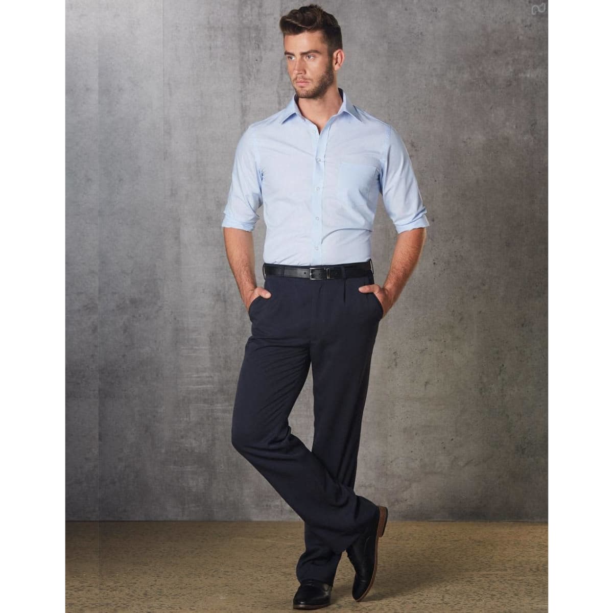 A man in a light blue shirt and black permanent press pants stands with hands in pockets against a grey backdrop.