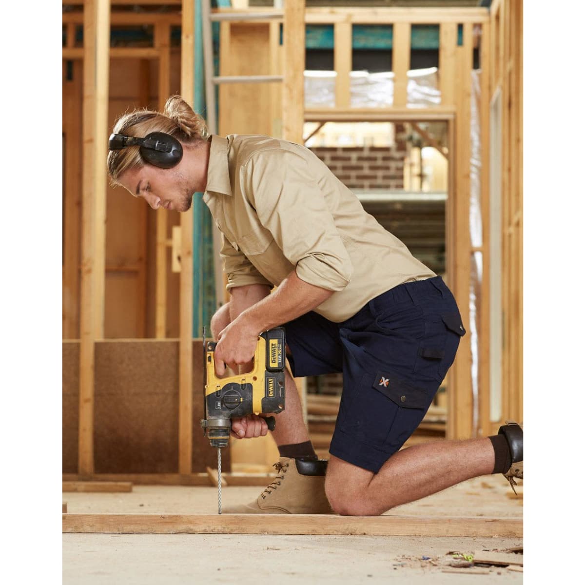 A man in khaki and navy work shorts operating a drill, wearing ear protection, in a construction setting.
