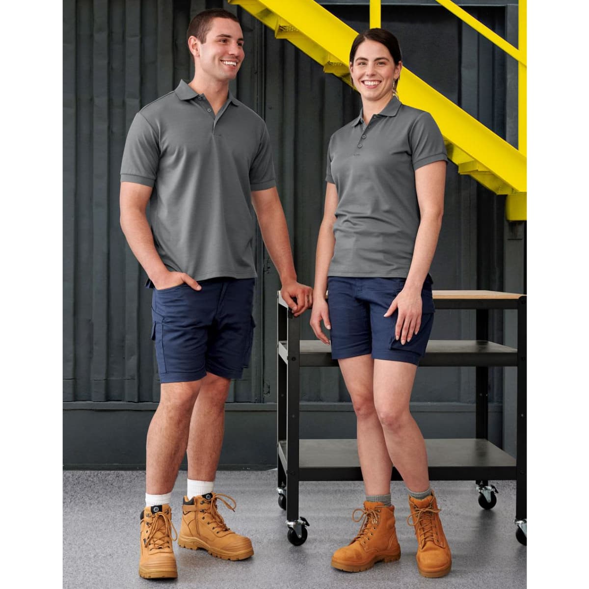 A pair of models wear unisex cotton stretch ripstop work shorts in navy, paired with grey polo shirts and work boots.