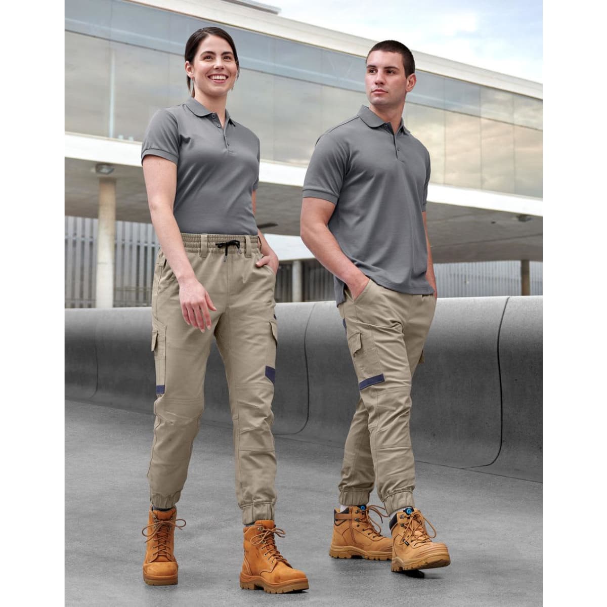 Unisex cotton stretch drill cuffed work pants in khaki, modelled by two people wearing grey tops and work boots.