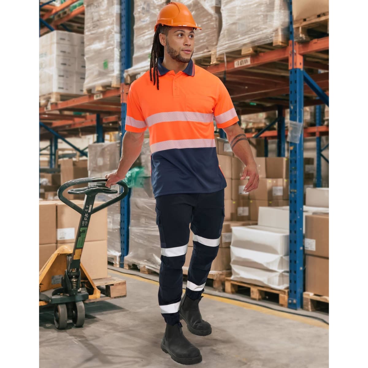 A man wearing orange and navy work pants with reflective tapes, accompanied by a hard hat and boots, stands in a warehouse.