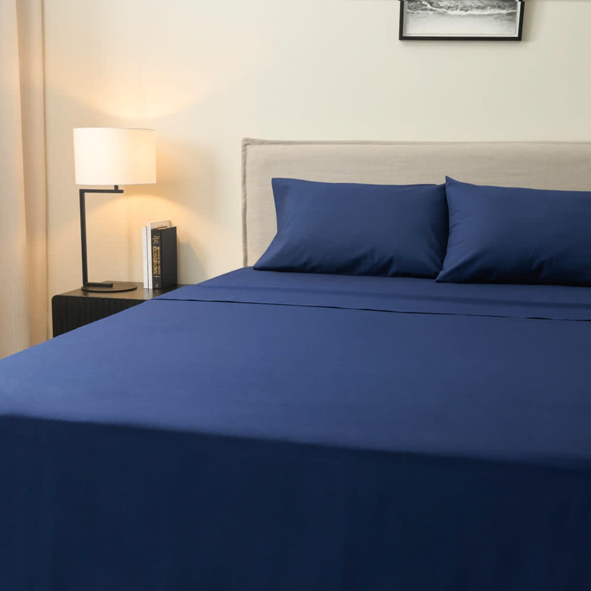 Navy blue vat dyed bed sheets neatly arranged on a bed with two matching pillowcases, beside a lamp and book.