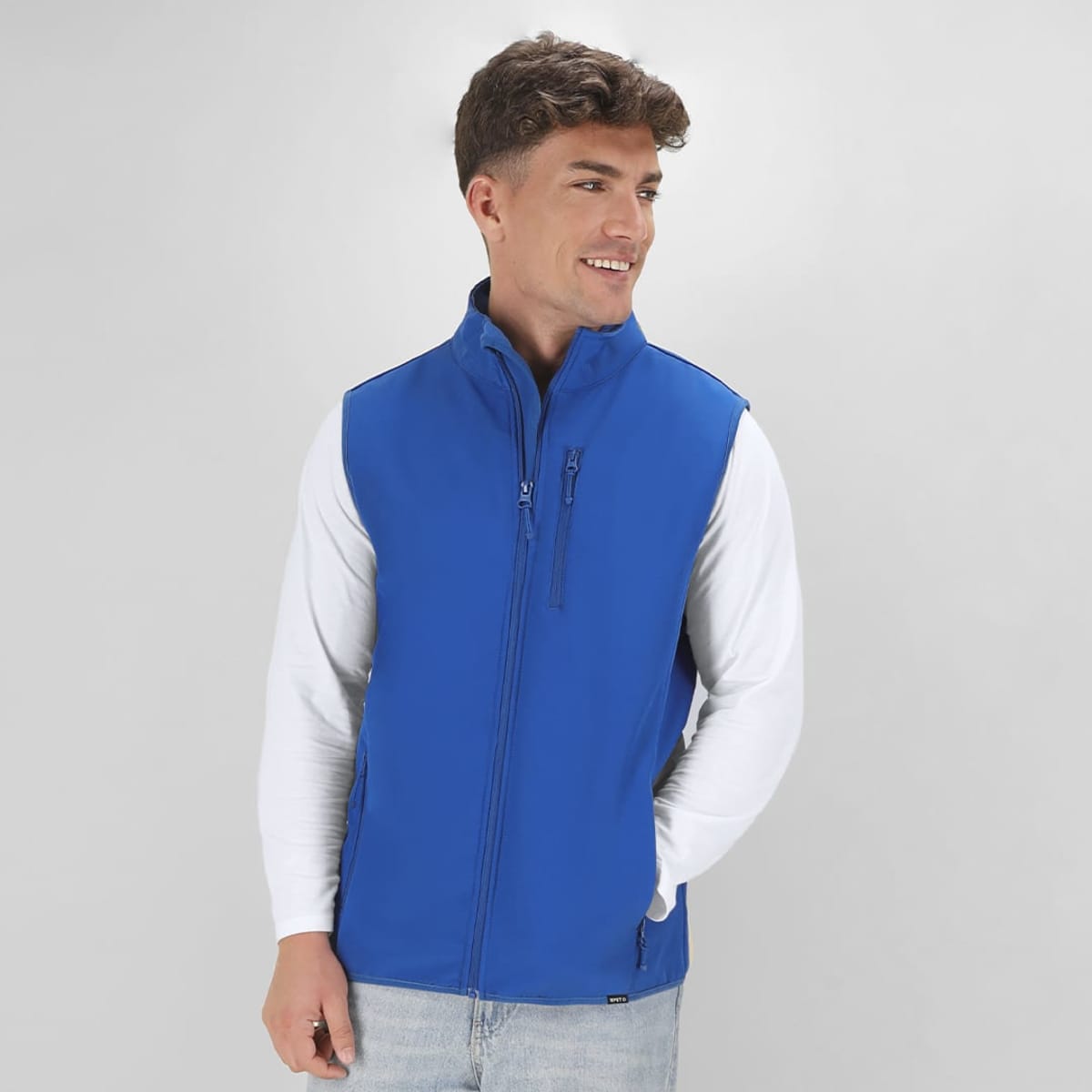 A blue vest jacket is worn by a smiling young man, featuring a zip front and a logo on the chest.