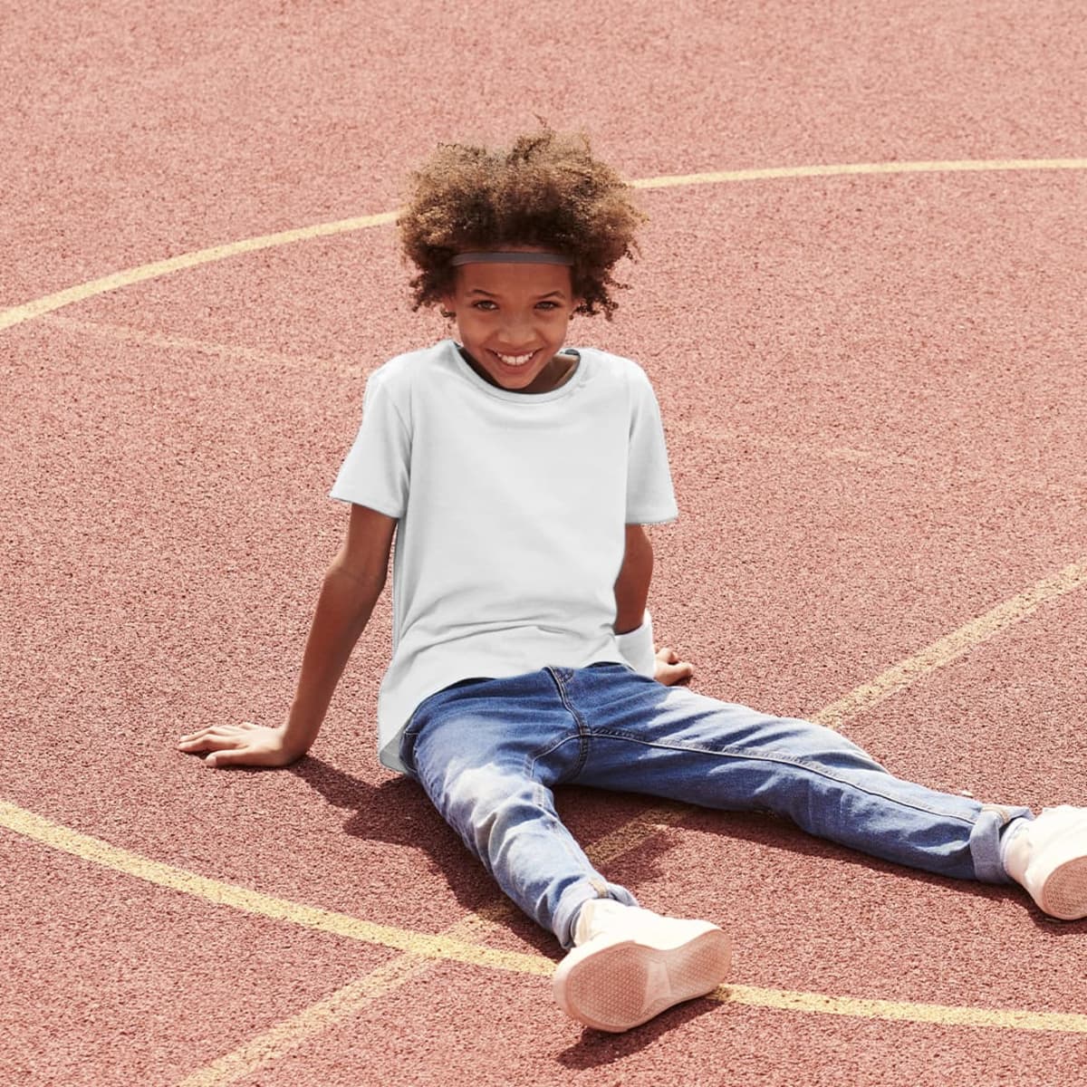 A child sits on a sports court wearing a light blue t-shirt and blue jeans, smiling at the camera.