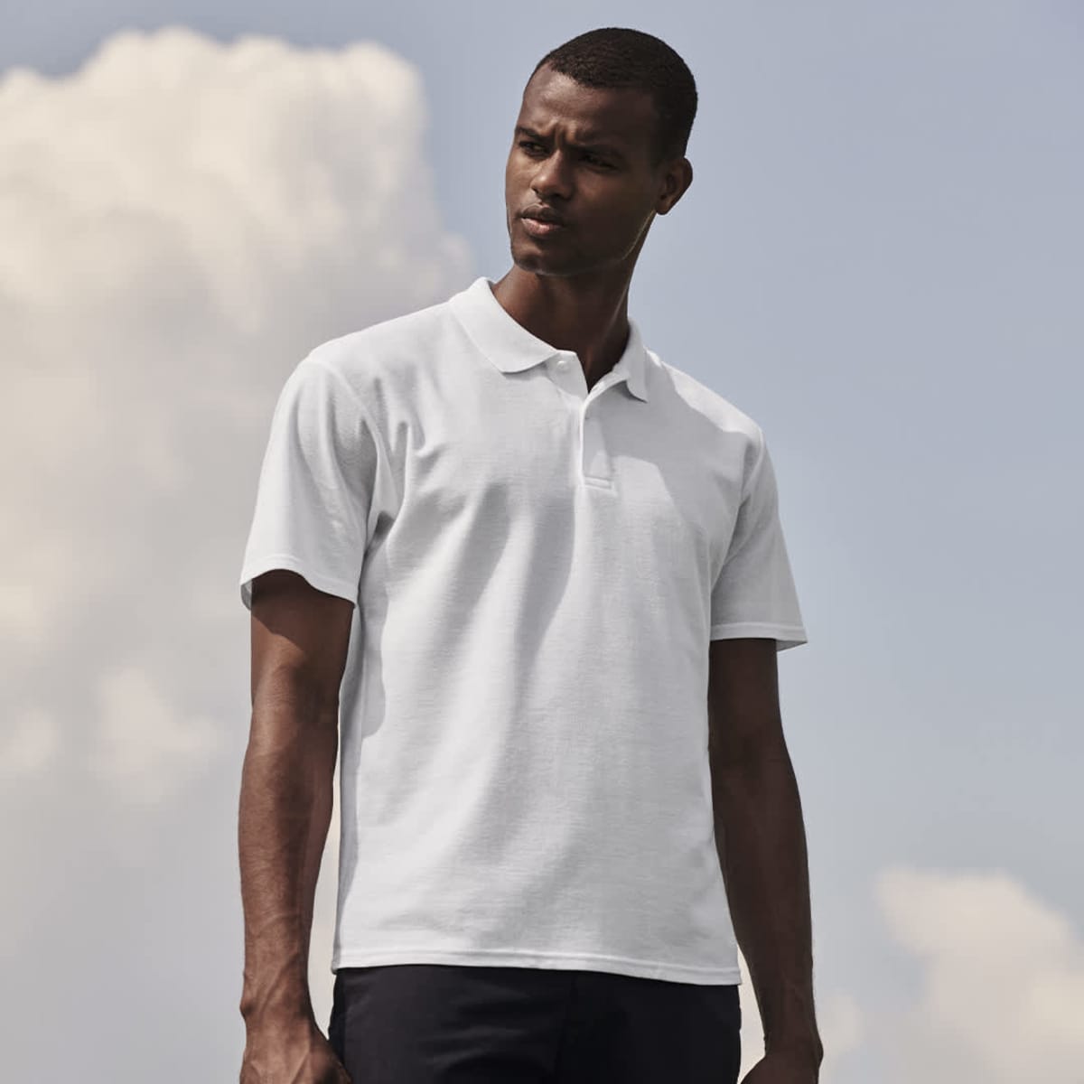 A white polo shirt on a model stands against a cloudy sky, featuring a simple design and a logo on the chest.