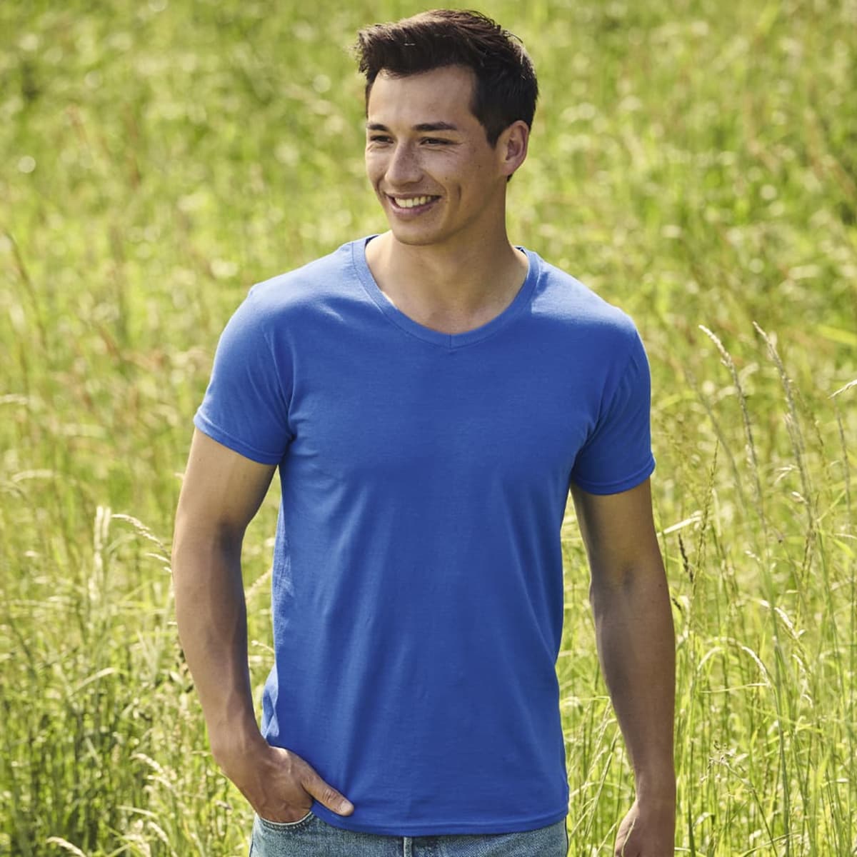 A man wearing a blue Iconic V-Neck T-Shirt stands in a grassy field. The shirt has a logo.