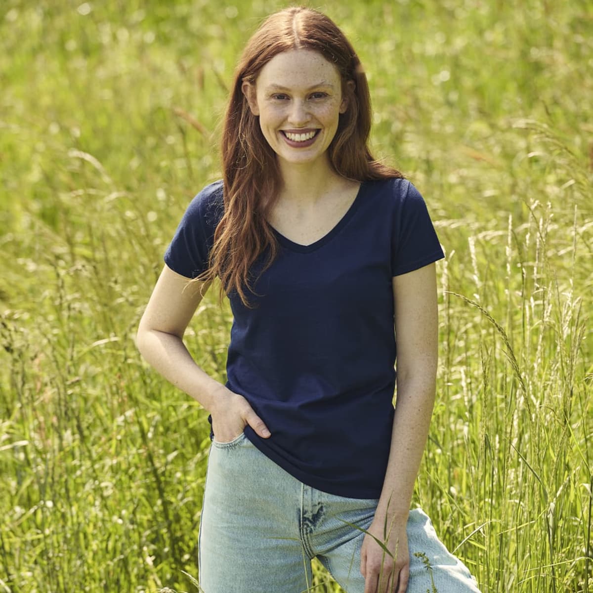 A young woman wearing a navy Iconic V-Neck T-Shirt stands in a grassy field, smiling while posing with her hand in her pocket.