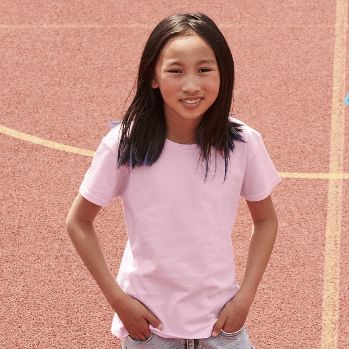 A young girl poses in a light pink t-shirt on a red sports court, with her hands in her pockets.