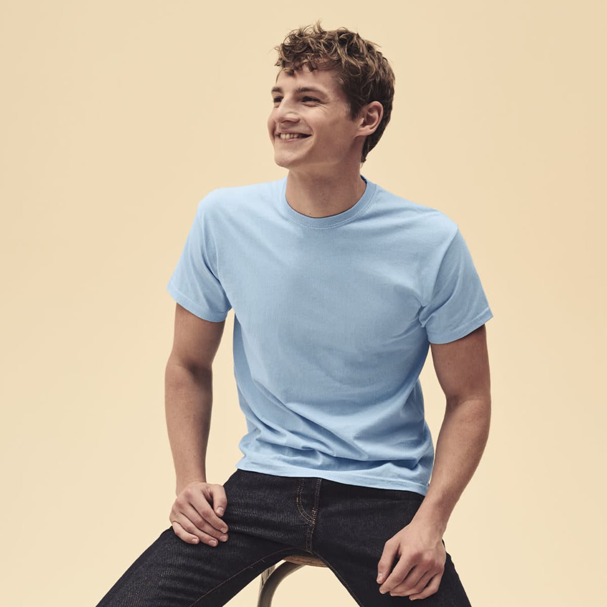 A light blue T-shirt worn by a smiling male model, seated on a stool against a neutral background.