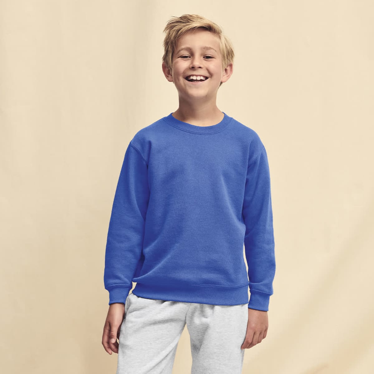 A blue sweat hoodie on a smiling child, paired with light grey trousers. The hoodie has a logo.