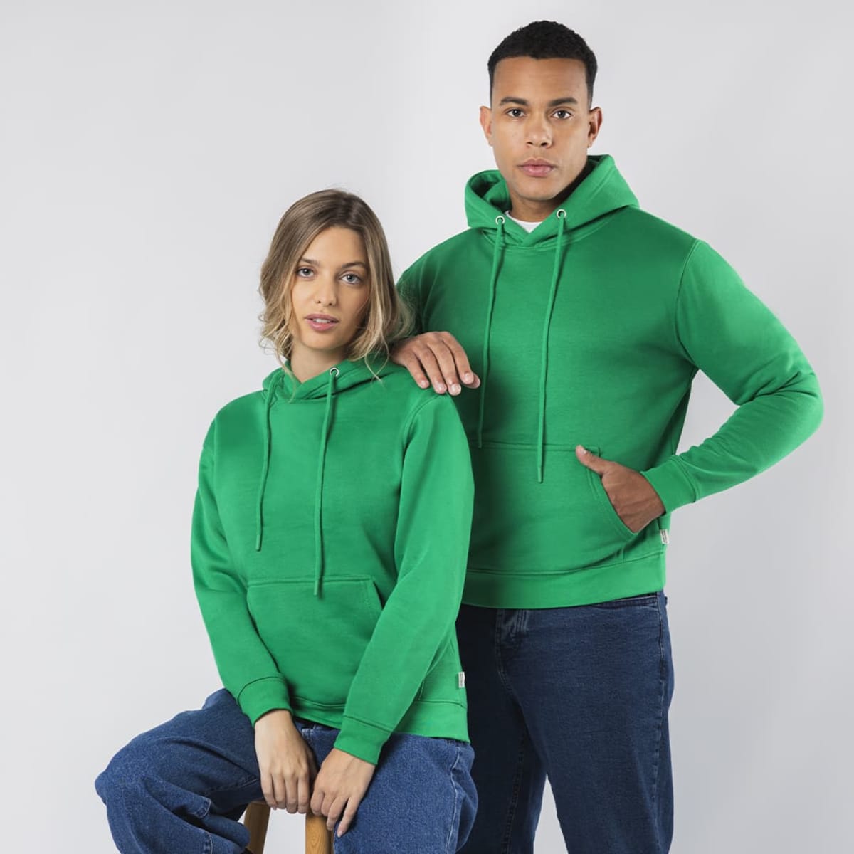 A green hoodie with a front pocket, displayed by a woman sitting and a man standing beside her against a plain background.