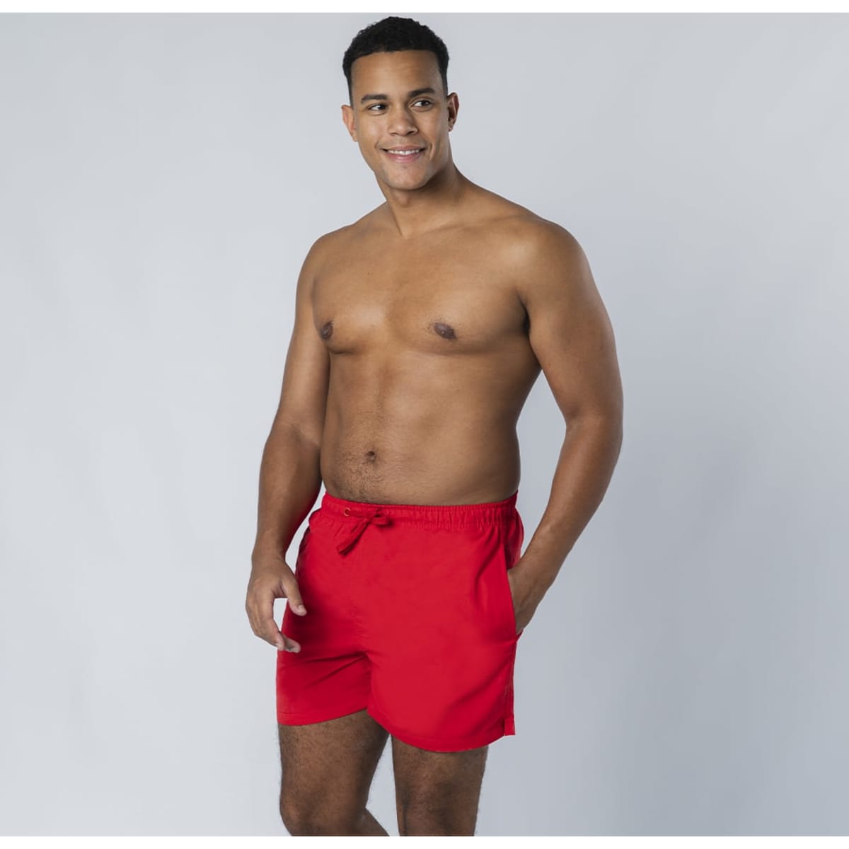A man with a toned physique is wearing red swim shorts, standing against a grey background.