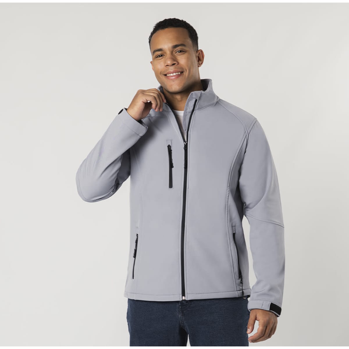 A light grey soft shell jacket is worn by a smiling person, featuring a small zippered pocket and a logo.