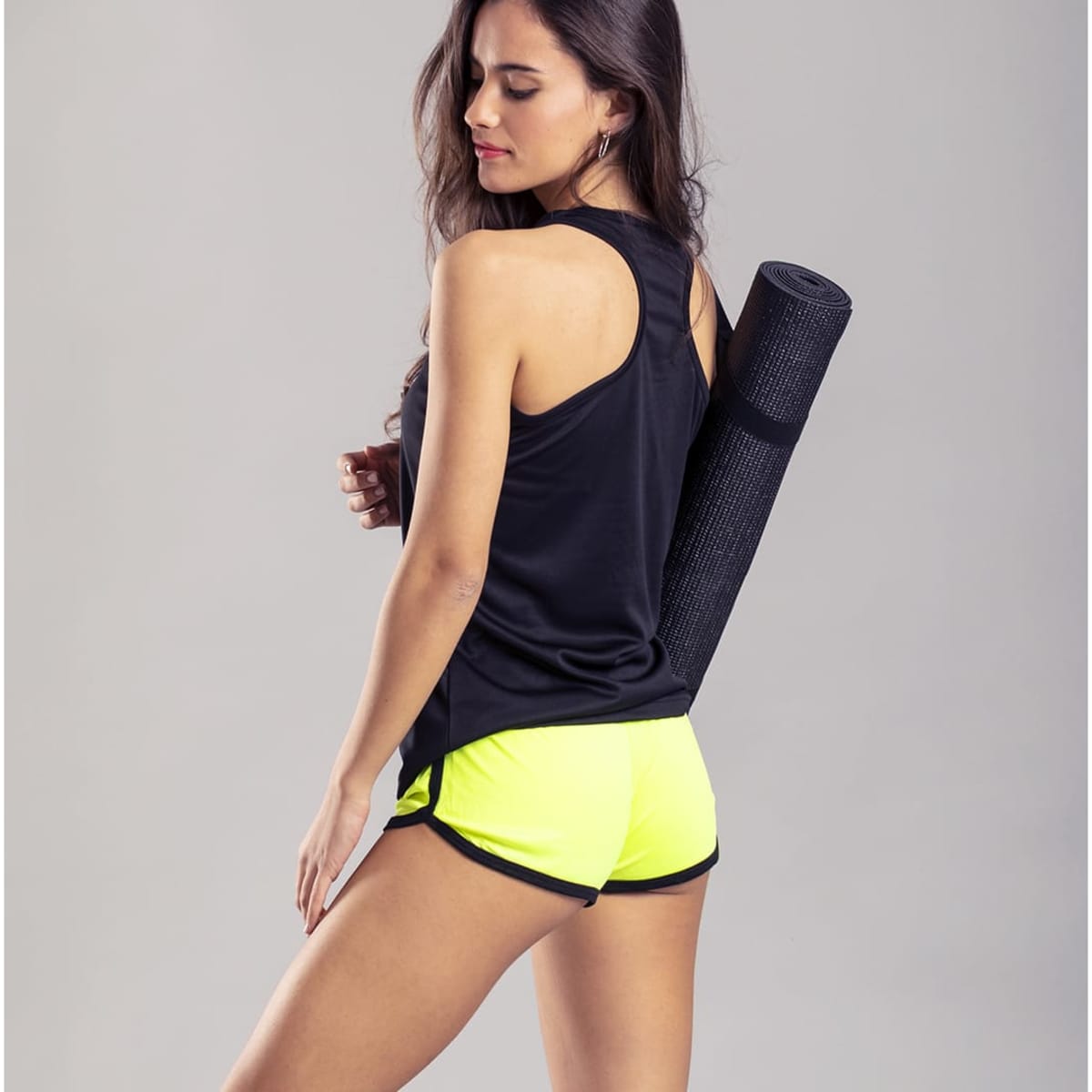 A woman is wearing black activewear and bright yellow dress shorts, holding a yoga mat. The outfit features a logo.