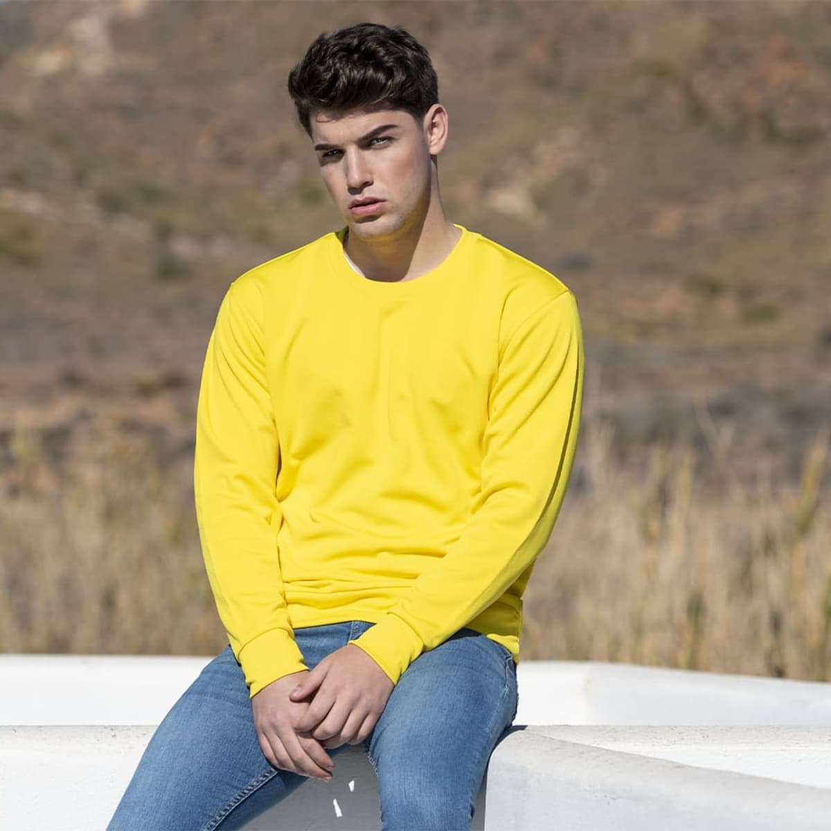 A young man wearing a bright yellow long-sleeve hoodie and blue jeans, sitting outdoors against a textured background.