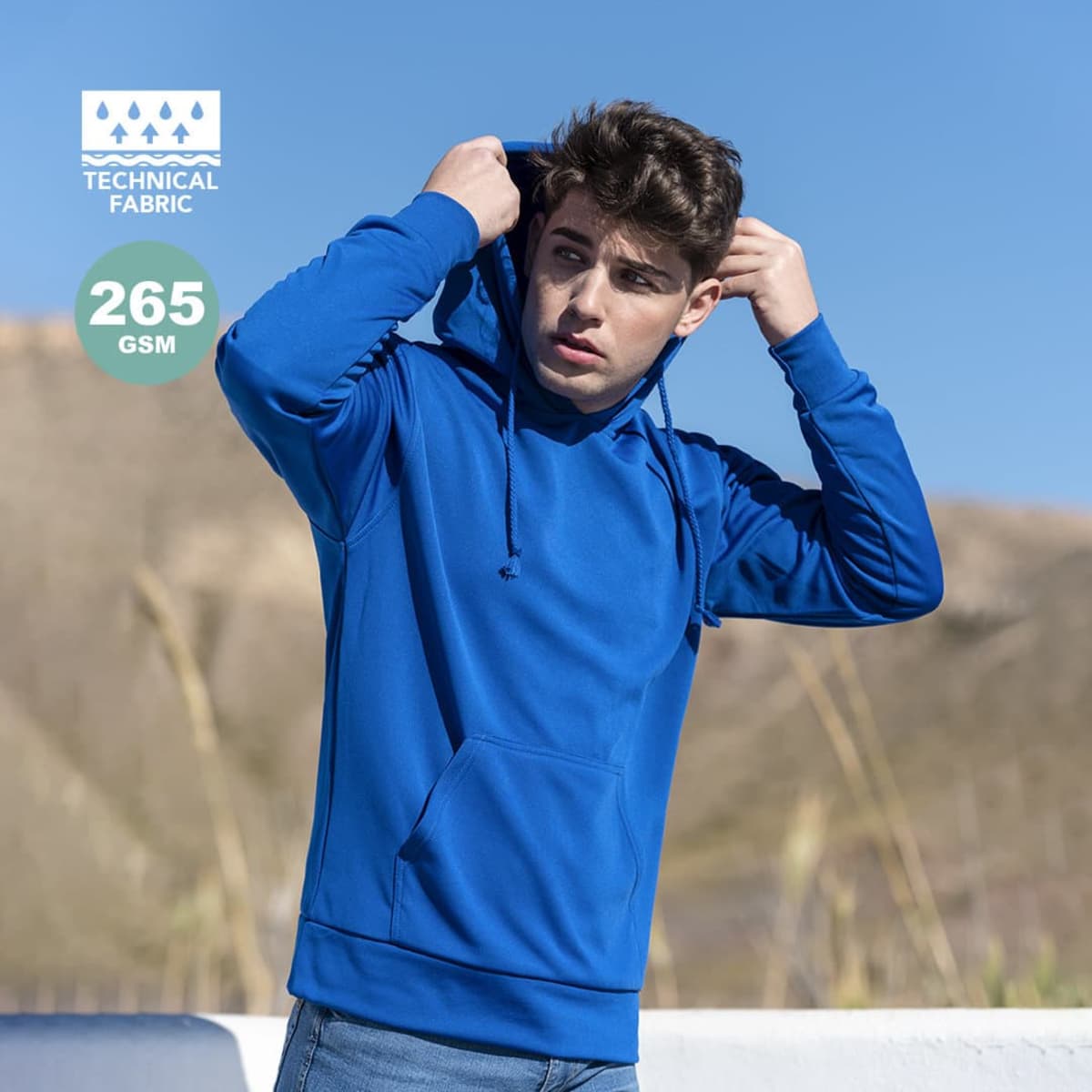 A blue technical sweatshirt with a hood, featuring a logo and worn by a person against a natural background.