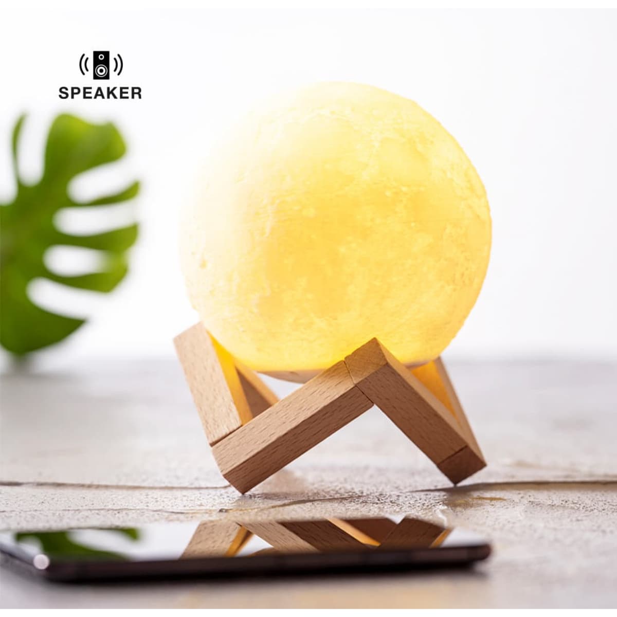 A round yellow moon-shaped speaker sits on a wooden stand beside a smartphone on a surface.