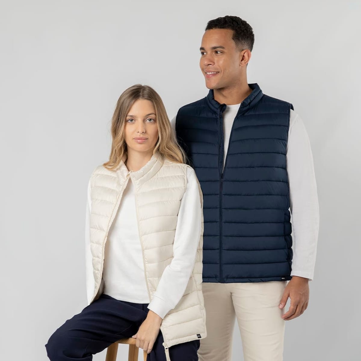 A cream vest and a navy vest are worn by two models against a neutral backdrop, both featuring a logo.