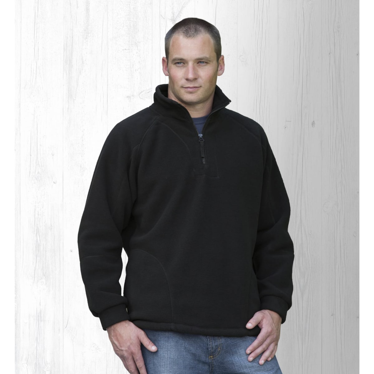 A man wearing a black polar fleece pullover with a half zip, standing against a light wooden background.