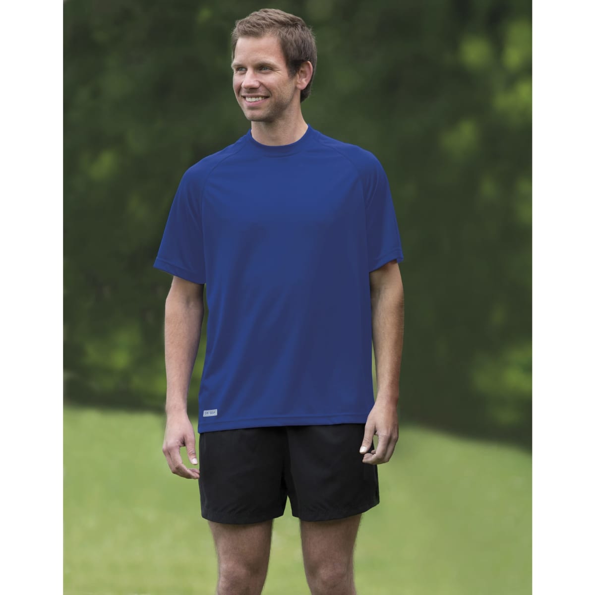 A man stands outdoors, wearing a blue Dri Gear Plain Raglan Tee and black shorts. The shirt has a logo on it.