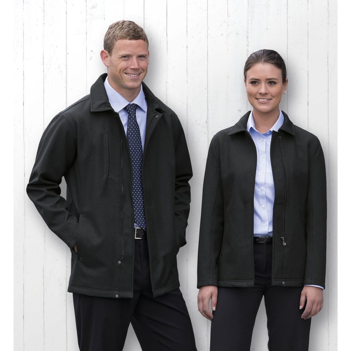 A men's and women's black soft shell jacket displayed on two models against a white wooden background. Each jacket features a logo.