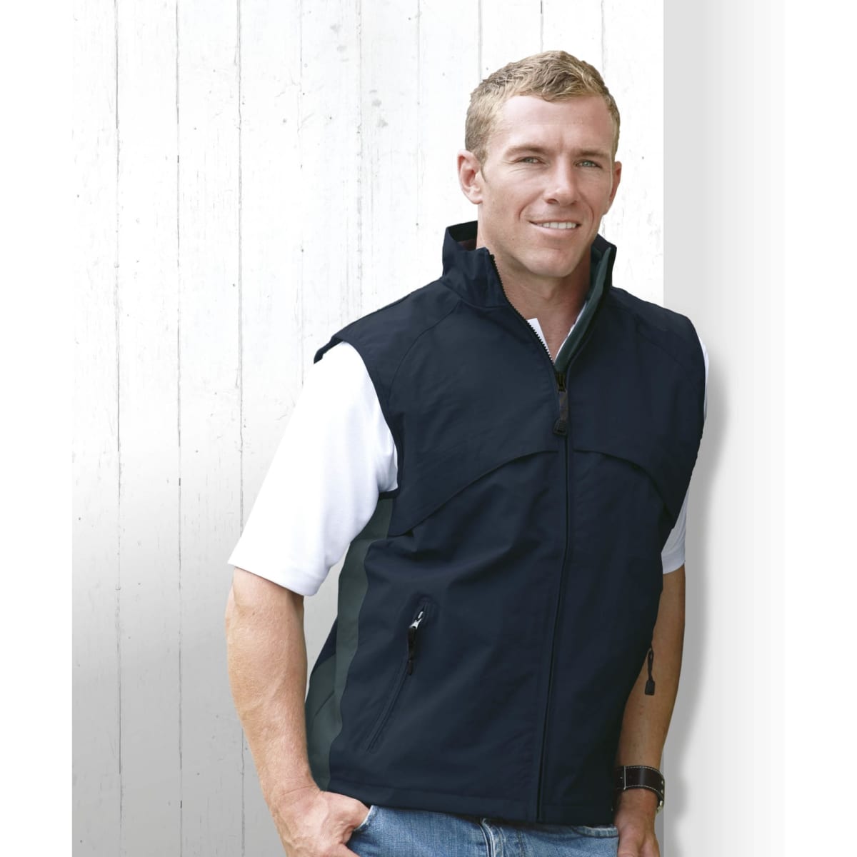 A man wearing a black vest, standing against a white wooden background. The vest features a zip and side pockets.