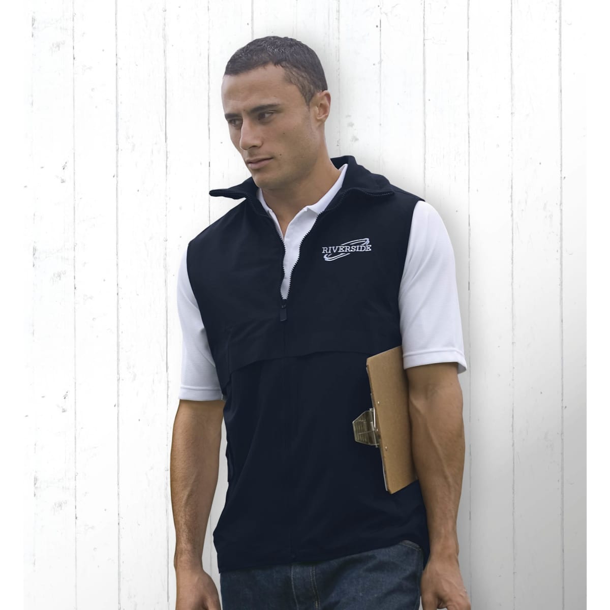 A man wearing a black nylon vest with a logo, holding a clipboard, standing against a white wooden background.