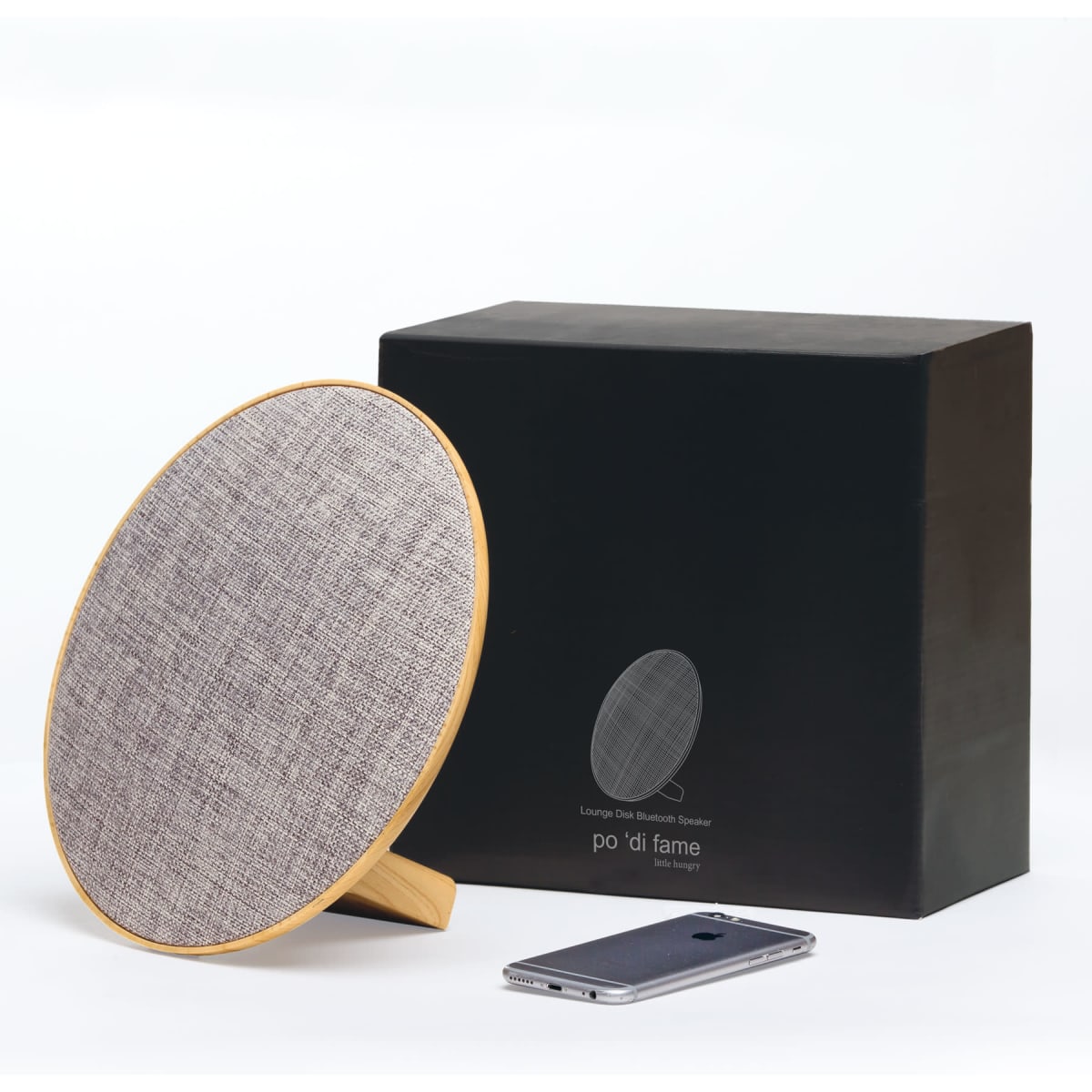 A round grey Bluetooth speaker with a wooden base next to a smartphone, accompanied by a black box packaging.