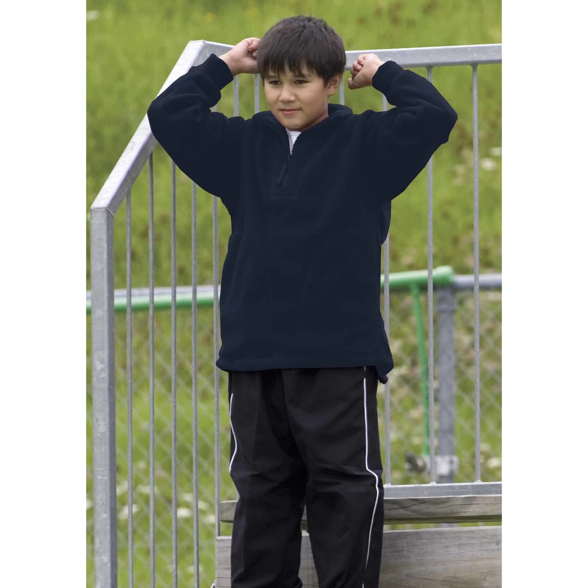A boy wearing a black detailed polar fleece pullover stands on a metal structure, with green grass in the background.