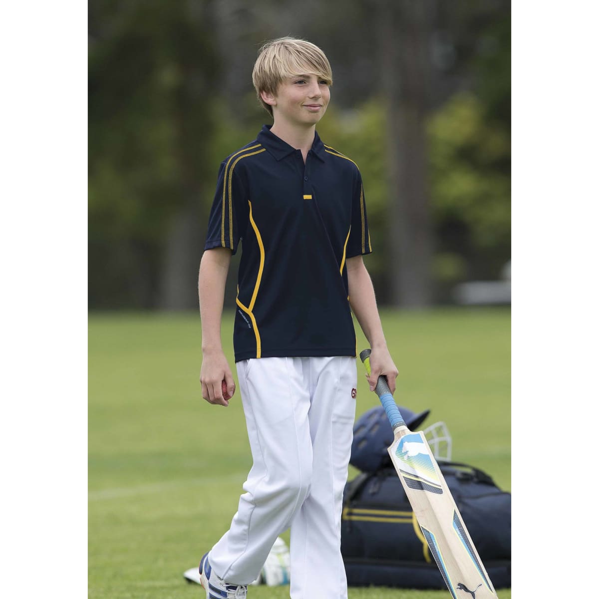 A youth wearing a navy polo shirt with yellow accents, walking on a cricket field with sporting equipment.