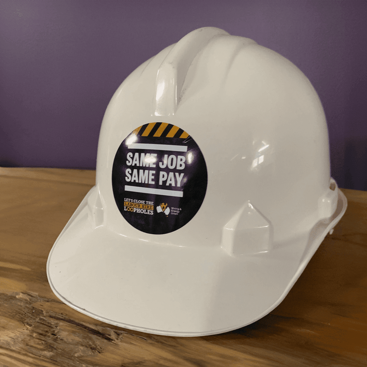 A white hard hat featuring a circular sticker. The sticker has a logo and bold text.
