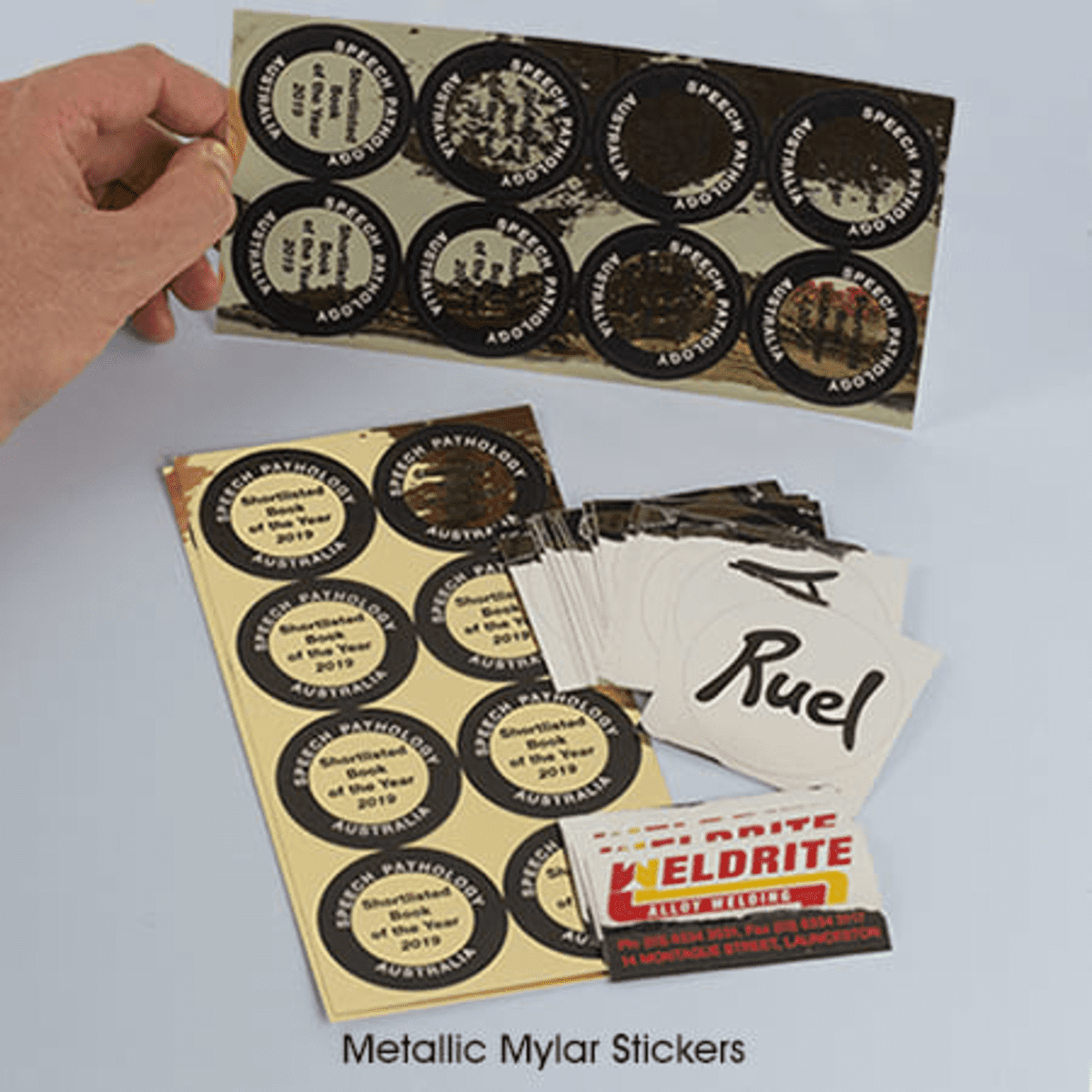 A hand holding a sheet of metallic Mylar stickers featuring circular and rectangular designs in black, white, and gold.