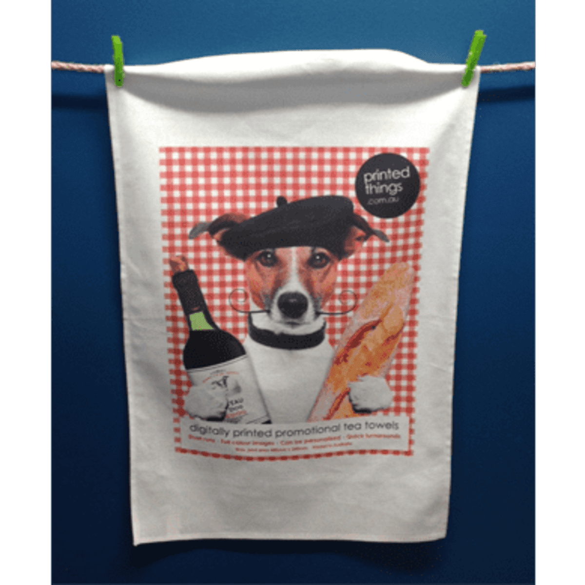 A white tea towel featuring a dog in a beret, holding a wine bottle and a baguette against a red checkered background.