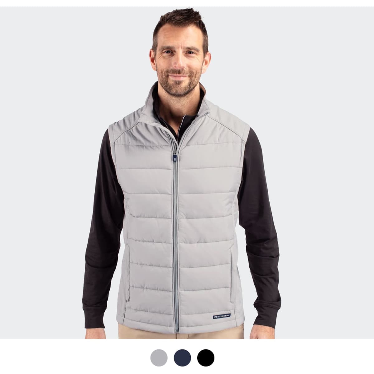 A man in a light grey quilted vest stands against a plain background. The vest features a branding logo.