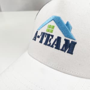 A white trucker cap featuring a blue and green logo with the text "A-Team" in navy embroidery.
