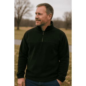 A man is wearing a black 1/2 zip polar fleece jacket, standing outdoors in a natural setting. The jacket has a logo.