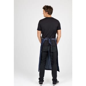 A man wearing a long waist cotton/denim apron in blue, with a black t-shirt and jeans, facing away from the camera.