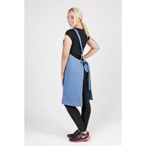 A woman wearing a denim apron, showcasing its full body design. The apron features a tie at the back and is a blue shade.